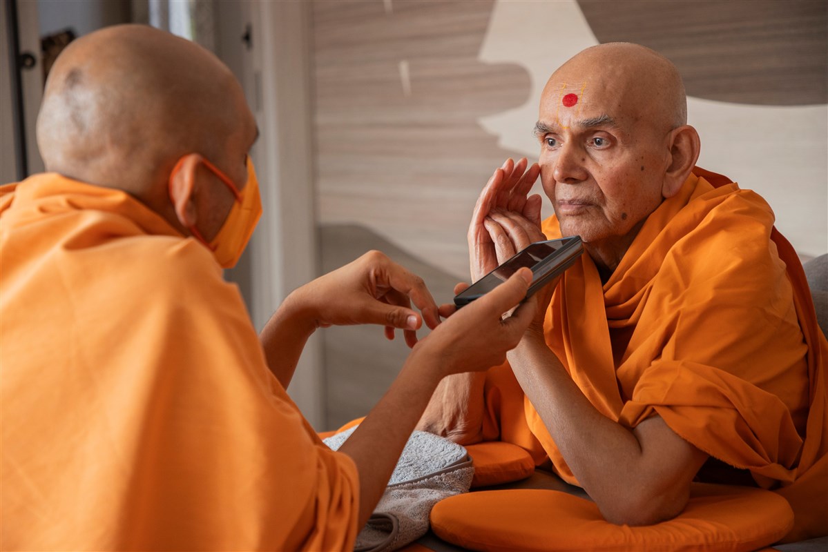 Param Pujya Mahant Swami Maharaj seated, hand raised to his ear, listening to a phone held out by a sadhu during vicharan at Robbinsville.